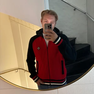 Röd och svart Cardgian från J.Lindeberg  - Säljer en snygg röd och svart cardigan från J.Lindeberg med dragkedja och huva. Tröjan har stickad känsla, svarta ärmar och detaljer i vitt och rött på muddar och nederkant. Två fickor med dragkedja och en liten logga på bröstet. Perfekt för höst och vinter! Litet hål på insidan av dragkedjan, priset går att diskutera 