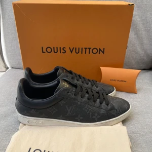 Louis Vuitton skor - Svarta Louis Vuitton sneakers i skinn med klassiskt monogrammönster över hela skon. Snygga svarta skosnören och vit sula med diskreta LV-detaljer på sidan. Perfekta för dig som vill ha en lyxig och stilren look med streetkänsla. Dustbag medföljer bara!