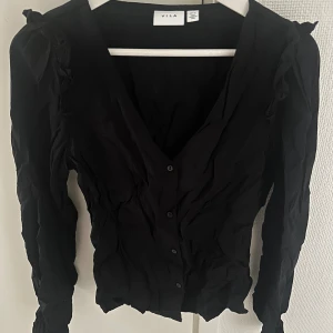 Svart blus med volang från Vila - Svart blus från Vila med v-ringning och knappar framtill. Långa ärmar och volangdetaljer på axlarna ger en extra touch. Jättebra skick, behöver strykas!