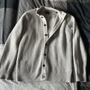 Grå ribbad kofta från Massimo Dutti - Snygg grå kofta från Massimo Dutti i ribbstickad ullmix. Modellen har lång ärm, knappar framtill och en klassisk krage. Perfekt för dig som gillar stilrena och tidlösa plagg med skön passform.