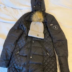 Svart quiltad dunjacka Rock and blue med mössa - Snygg svart lång quiltad dunjacka från Rockandblue med huva och dragkedjor på fickorna. Jackan har ett stilrent mönster och Rockandblue-logga på ärmen. Perfekt för kalla dagar och ger en cool vibe till vinterlooken.