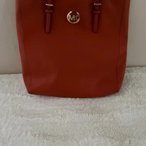 Orange axelväska från Michael Kors - Snygg orange axelväska från Michael Kors i slätt skinn med guldfärgat MK-emblem framtill. Väskan har raka linjer, två långa axelremmar och rymligt innerfack. Perfekt för dig som vill ha plats för allt och samtidigt hålla stilen.