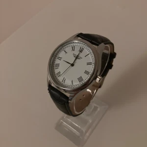 Klassisk BINLIHUAN klocka med svart läderband - Stilren klocka från BINLIHUAN med vit urtavla, svarta romerska siffror och silverfärgat boett. Klockan har ett svart läderband med krokodilstruktur och klassiskt spänne.                                                                                 ✅Helt Ny✨.                                                                                    ✅Frakt inom 24 timmar📦.                                                      ✅Kommer med Låda 🗃️.