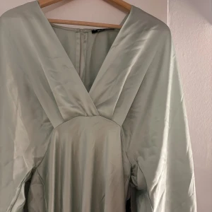 Ljusgrön långklänning i satin - Elegant ljusgrön långklänning från Gina Tricot i glansigt satinmaterial. Klänningen har djup v-ringning, vida långa ärmar med resår vid handlederna och en snygg siluett som faller mjukt längs kroppen. Perfekt som balklänning eller liknande. Aldrig använd, nyskick☺️