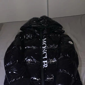 Svart glansig dunjacka från Moncler - Svart glansig dunjacka från Moncler med stor huva och tydlig logga på ärmen. Jackan har puffig design, dragkedja framtill och stora vita bokstäver längs dragkedjan. Perfekt för kalla dagar och riktigt snygg streetstil.
