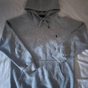 Grå hoodie från Polo Ralph Lauren - Snygg grå hoodie från Polo Ralph Lauren med klassisk broderad logga på bröstet. Tröjan har huva med snörning och en stor magficka. Perfekt för chill dagar och enkel att matcha med jeans eller joggers. Storlek M men passar även S 