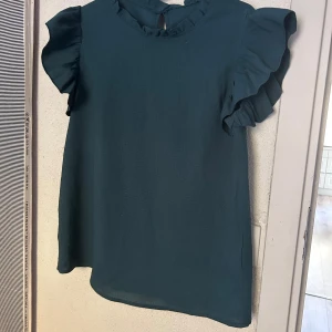 Mörkgrön blus med volangärmar - Supersöt mörkgrön blus med volangdetaljer på ärmarna och liten volang vid halsringningen. Blusen har en rak passform och knäpps med en knapp i nacken. Perfekt för dig som gillar en feminin och trendig stil.