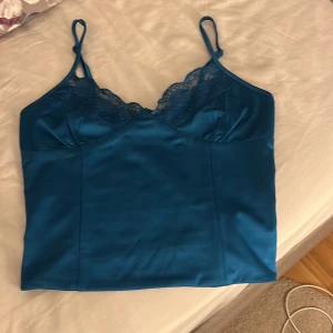 Blå satin linnetopp från BikBok - Säljer en blå linnetopp från BikBok i satinliknande material med spetsdetaljer vid bysten och smala justerbara axelband. Toppen har en slät och glansig finish. Säljer då den inte kommer till användning 