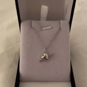 Halsband med hjärta och pil i silver liten - Säljer ett snyggt halsband i silverfärg med ett hjärta som har en pil genom sig. Kedjan är tunn och smycket har en modern, stilren look. Perfekt accessoar för att lyfta din outfit. Rostfri