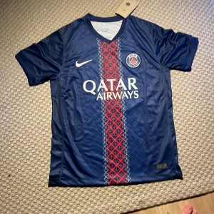 PSG Dembele matchtröja Nike S - Säljer en officiell Paris Saint-Germain fotbollströja från Nike med O. Dembélé och nummer 10 på ryggen. Tröjan är marinblå med röd och vit rand i mitten, PSG-logga och Qatar Airways-tryck på bröstet. Tillverkad i lätt och ventilerande material, perfekt för fotbollsplanen.