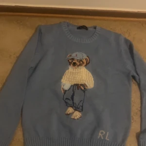Blå stickad tröja Polo Ralph Lauren - Säljer en ljusblå stickad tröja från Polo Ralph Lauren med ikonisk björn på bröstet. Tröjan har rund halsringning, långa ärmar och ribbade muddar. RL-broderi nertill. Perfekt för dig som gillar klassisk preppy stil.