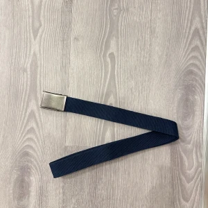 Mörkblå canvasbälte med metallspänne - Snyggt mörkblått bälte i slitstark canvas med ett stilrent, rektangulärt metallspänne i silverfärg. Bältet har en enkel och clean design som passar till jeans eller chinos. Perfekt för dig som gillar minimalistiska accessoarer och vill ha något som funkar till många olika outfits.