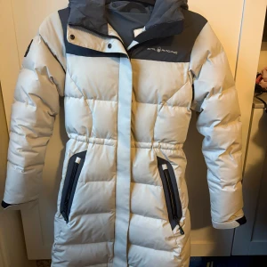 Vit och ljusgrå dunjacka Sail Racing - Intresse koll på min långa och riktigt varm dunjacka från Sail Racing i vit med mörkgrå detaljer. Jackan har huva, två stora fickor med dragkedja och snygga logopatchar på ärm och bröst. Perfekt för kalla vinterdagar och har en clean, sportig look.