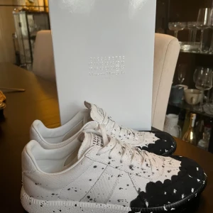 Maison Margiela GATS - Maison Margiela GATS sneakers i vit canvas med svart och vit färgstänk-design. Skorna har snörning, rund tå och platt sula. Märkets ikoniska etikett syns på plösen. Unik och edgy look som sticker ut i mängden. Storlek 43😊
