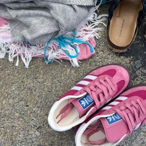 Adidas Gazelle rosa sneakers strl 37/38 - Säljer ett par Adidas Gazelle sneakers i en snygg rosa färg med vita ränder och detaljer. Skorna har klassisk låg profil, rund tå och platt sula. Ovandelen är i mocka och textil, med snörning framtill och vit insida. Helt nya och oanvända!!!