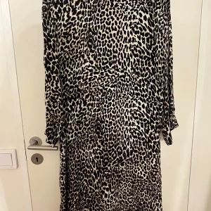 Leopardmönstrad långklänning från Fiore - Säljer en snygg långklänning från Fiore med leopardmönster i svart och beige. Klänningen har långa ärmar och en avslappnad passform. Perfekt för dig som vill sticka ut med ett djurmönstrat plagg!