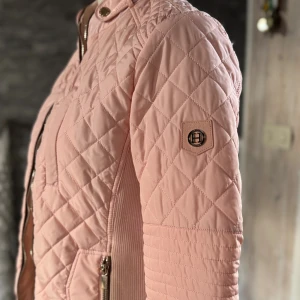 Ljusrosa quiltad jacka från Hollies - Säljer en ljusrosa quiltad jacka från Hollies i storlek 38. Jackan har snygga detaljer som dragkedjor i guld, ståkrage med knappar och ribbade partier i sidorna. Perfekt för vår och höst när du vill ha en stilren look.