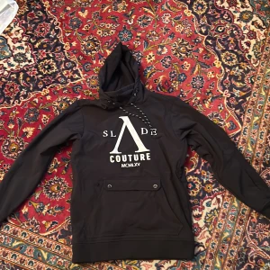 Svart hoodie från Slade Couture S - Svart hoodie från Slade Couture med stor vit logga framtill och mindre tryck på ryggen. Tröjan har huva med snörning, långärmade ärmar och en praktisk ficka med knappar på magen. Materialet känns som polyester och insidan är mjuk. Snygg och streetig stil. Har luft fickor vid sidan.