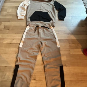 Beige mjukisbyxor från Kappahl - Snygga beige mjukisbyxor från Kappahl i storlek 140. Byxorna har svarta och vita detaljer längs sidorna och elastisk midja med snörning. Perfekta för chill dagar och har en loose passform som ger skön rörelsefrihet.