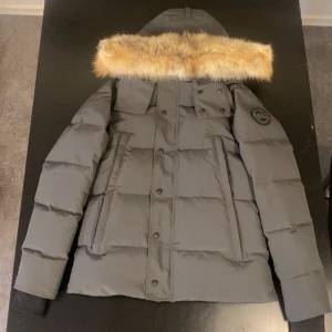 Grå dunjacka med fuskpäls från Canada Goose - Snygg grå dunjacka från Canada Goose med avtagbar huva och fuskpäls. Jackan har flera fickor framtill, ribbade muddar och klassisk patch på ärmen. Perfekt för kalla vinterdagar och riktigt varm. Stängs med både dragkedja och knappar. Storleken är xs men passar också s.