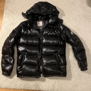 Svart Moncler pufferjacka med huva - Svart pufferjacka från Moncler med glansig finish, huva och dragkedja framtill. Jackan har två snedställda fickor med dragkedja och Moncler-logga på ärmen. Insidan har ett unikt serietidningsmärke. Perfekt för kalla dagar och riktigt snygg streetstil.