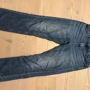 Jack & Jones blå jeans Regular Clark - Snygga blå jeans från Jack & Jones, modell Regular Clark. Klassisk femficksdesign med ljusare tvättade partier och kontrastsömmar. Rak passform och normal midja, tillverkade i slitstarkt denim. Perfekta för dig som gillar en enkel och tidlös jeansstil.