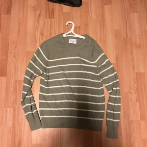 Stickad tröja från Vailent  - Snygg stickad tröja från Vailent i storlek XS. Den är olivgrön med vita horisontella ränder och har en rund halsringning. Tröjan är ribbstickad och har långa ärmar, perfekt för att hålla dig varm och stilren under kyliga dagar.