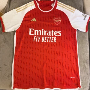 Arsenal hemmatröja 2023/24 Adidas - Säljer en officiell Arsenal hemmatröja från säsongen 2023/24, tillverkad av Adidas. Tröjan är röd med vita korta ärmar, guldiga detaljer och blixtmönster över hela framsidan. Materialet är lätt och andas, perfekt för match eller träning. Emirates Fly Better-logga på bröstet.