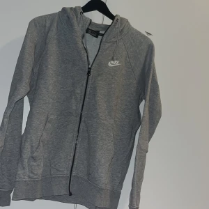 Grå hoodie från Nike med dragkedja - Grå Nike hoodie med hel dragkedja framtill och klassisk huva. Tröjan har långa ärmar, två fickor och en liten vit Nike-logga på bröstet. Perfekt för chill dagar eller när du vill ha en sportig look.