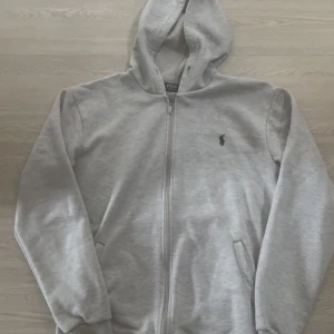 Grå hoodie från Polo Ralph Lauren - Snygg ljusgrå hoodie från Polo Ralph Lauren med dragkedja och klassisk broderad logga på bröstet. Tröjan har huva, fickor framtill och är fodrad med mjuk fleece på insidan. Perfekt för chill dagar och enkel att matcha med jeans eller joggers.