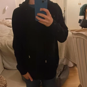 Svart hoodie i merinoull  - Svart hoodie med klassisk huva och snörning framtill. Tröjan har långärmad design och en loose passform som ger en chill vibe. Perfekt för att slänga på sig när det är lite kyligt ute. Enkel och stilren utan synliga loggor eller mönster.