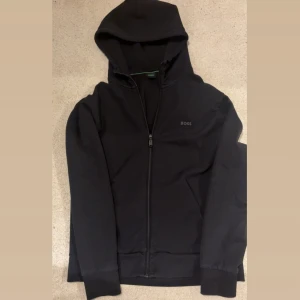 Svart Hugo BOSS kofta - Svart hoodie från BOSS med logga på bröstet. Tröjan har huva, hel dragkedja. Perfekt för chill dagar eller när du vill ha en clean look. Storlek är L. Oanvänd, kvitto finns.