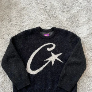 Svart stickad tröja från Corteiz - Svart stickad tröja från Corteiz med stor vit C och stjärna på framsidan. Rund hals och långa ärmar, ribbade muddar. Materialmix av mohair, ull, nylon och akryl som ger en fluffig och mjuk känsla. Perfekt för dig som gillar streetwear med unik design.
