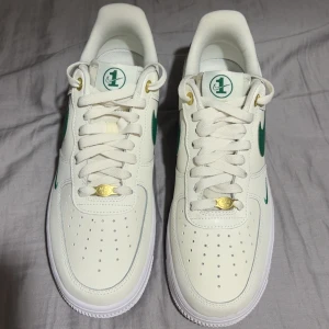 Nike Air Force 1 vit/grön sneakers - Helt nya aldrig använda Nike air force! Nike Air Force 1 sneakers i vitt skinn med gröna detaljer och logga. Klassisk låg modell med perforerad tå, grönt swoosh på sidan och guldiga detaljer vid snörningen. Vit sula med gröna cirklar undertill och AIR-tryck på sidan. Perfekta för en clean och sportig look. 