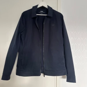 Mörkblå Nano Jacket från UBR - Snygg mörkblå Nano Jacket från UBR med stilren design och diskret logga på bröstet. Jackan har dragkedja framtill, klassisk krage och långa ärmar. Materialet är syntet med stretch och ventilerande egenskaper, samt avdunstar den vatten vid regn. Ny pris 3999:-