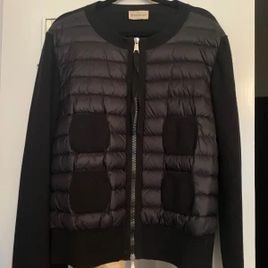 Svart pufferjacka från Moncler - Snygg svart pufferjacka från Moncler med quiltad framsida och stickade ärmar. Jackan har rund halsringning, dragkedja framtill och coola svarta detaljer på bröstet. Perfekt för dig som vill ha en stilren och trendig look.