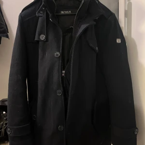 Mörkblå rock från Indicode Jeans - Snygg mörkblå rock från Indicode Jeans med klassisk krage, knappar framtill och dragkedja. Jackan har flera fickor med lock och knappdetaljer på axlar och ärmslut. Perfekt för dig som vill ha en stilren och tidlös look.