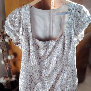 Designer kortklänning av Tiger of Sweden - Supersöt kortklänning från Tiger of Sweden med diskret skimrander mönster i beige, silver guld och vitt. Klänningen har puffiga korta ärmar, fyrkantig urringning och dragkedja bak. Tillverkad i ett glansigt, mjukt material som känns lyxigt och faller snyggt. Finns en fläck på insidan som ej går bort i tvätt. 