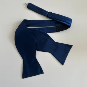 Marinblå fluga - helt ny - Klassisk gedigen marinblå (oknuten) fluga med justerbar halsrem och spänne, i len satin. Helt ny i förpackning.   Hämtas i Stockholm eller skickas mot porto. 