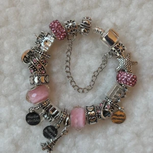 Charmarmband med rosa och silverdetaljer - Unikt charmarmband med flera berlocker i silverfärg och rosa. Armbandet har glittriga pärlor, hjärtan, stjärna, Eiffeltorn och textberlocker. Kedja i metall och detaljerade mönster gör det extra personligt och snyggt. Perfekt för dig som gillar att sticka ut med accessoarer.