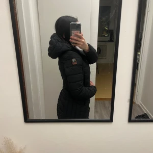 Svart dunjacka Parajumpers XXS - Svart, lång och lätt dunjacka från Parajumpers med huva och quiltad design. Jackan har dragkedja framtill, patch på ärmen och innerficka märkt 'Security Pocket'. Perfekt för kalla dagar och riktigt snygg passform.