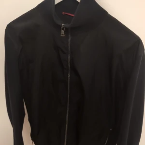 Svart prada Cardigan nyskick 10/10 - Svart bomberjacka från Prada med klassisk ribbad krage och ärmslut. Jackan har dragkedja framtill och diskret röd Prada-logga på ärmen. Materialmix med glansig framsida och stickade partier på ärmar och krage ger en clean och modern look.
