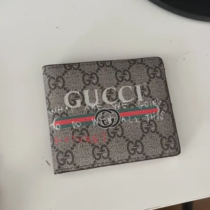 Gucci plånbok med GG-mönster - Snygg plånbok från Gucci med klassiskt GG-monogram i grått och brunt. Framsidan har texten 'What are we going to do with all this future?' och den ikoniska röd-gröna randen. Plånboken är i skinn och har flera kortfack samt fönster för ID. Varan är oanvänd, äkta och vid kort varsel kan priset gå ner