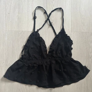 Svart  linnetopp med volang - Supersöt svart linnetopp med tunna korsade axelband! Toppen har små  broderier över hela tyget och är lätt utsvängd nedtill. Använd fåtal gånger och i topp skick! 