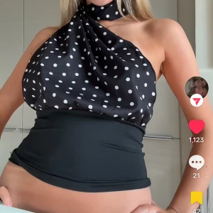Svart prickig topp - Säljer en svart topp med vita prickar och omlott-detalj. Toppen har en halterneck med knyt runt halsen och är axelbandslös på ena sidan. Materialet är glansigt och känns som satin, vilket ger en lyxig vibe. Perfekt för dig som vill sticka ut med en unik och trendig look.