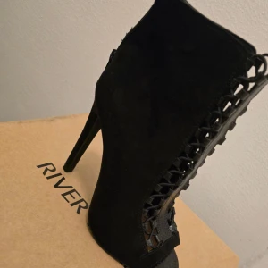 Svarta stövletter med klack från River Island - Snygga svarta stövletter från River Island i mocka-look med höga klackar och spetsig tå. Sidorna har coola utskurna detaljer med flätad design som ger en edgy vibe. Perfekta för dig som vill sticka ut med din stil.