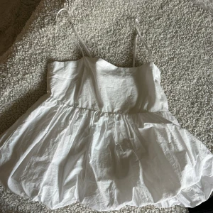 Vit volangig linnetopp från Pull&Bear - Supersöt vit linnetopp från Pull&Bear med tunna axelband och volangig, puffig nederdel. Toppen har en luftig och flowy passform som ger en riktigt fräsch vibe. Perfekt för varma dagar när du vill ha något lätt och stilrent.