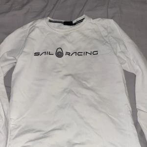 Vit långärmad tröja Sail Racing - Vit långärmad tröja från Sail Racing med svart logga tryckt över bröstet. Tröjan har rund halsringning med svart och vit randig detalj på insidan av kragen. Perfekt för en clean och sportig stil.