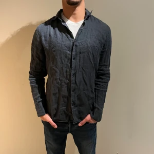 Mörkblå linneskjorta från Riley - Säljer en mörkblå långärmad skjorta från Riley i slim fit-modell. Skjortan är tillverkad i linne, har klassisk krage och knäppning framtill. Perfekt för dig som gillar en stilren och avslappnad look.