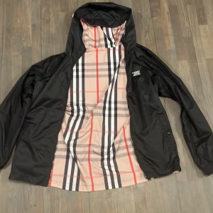 Burberry vindjacka med rutig design - Snygg vindjacka från Burberry med klassiskt rutig mönster i beige, svart, vitt och rött. Jackan har huva, dragkedja framtill och logga på bröstet. Perfekt för dig som vill sticka ut med en ikonisk streetstyle-look.